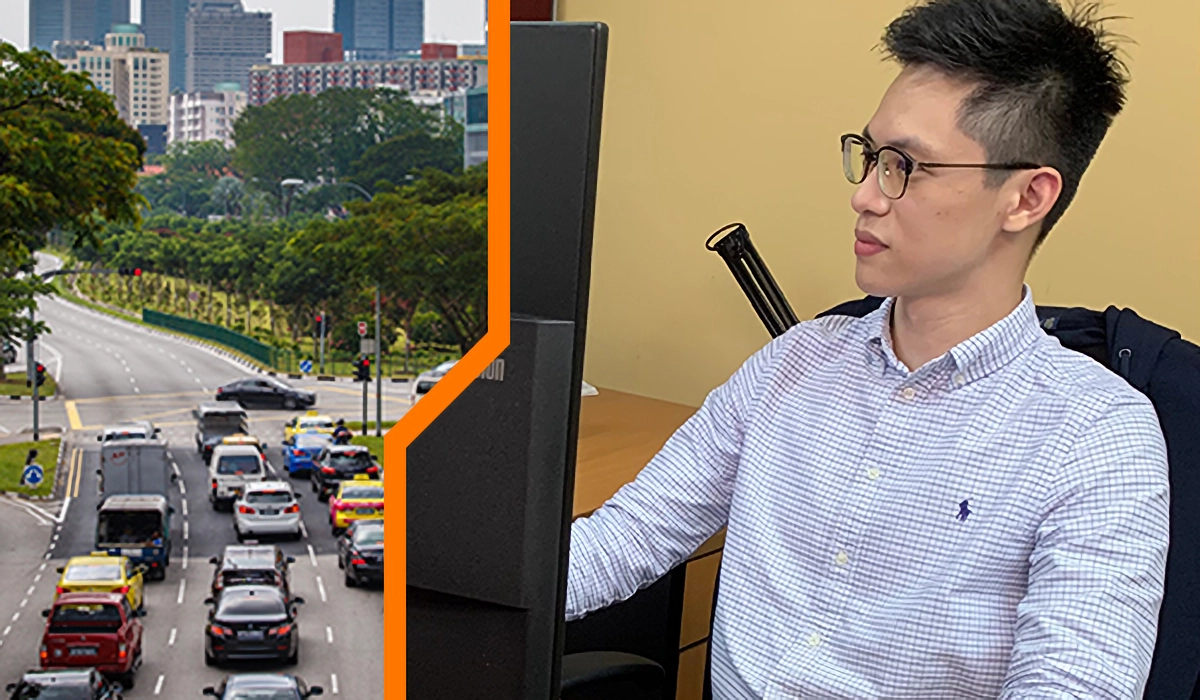 A young man with glasses sits at a desk beside a monitor, next to a city road with heavy traffic and tall buildings.